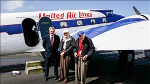Three white men stand in front of a small blue and white airplane. Two of the men are elderly.